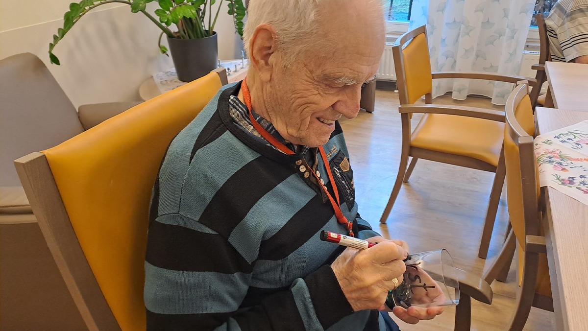 An older man sits in a cosy room, holding a personalised drinking glass and smiling contentedly. The homely environment with plants and furniture conveys peace and well-being.
