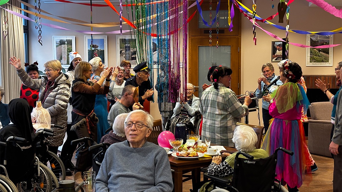 Residents in wheelchairs or dancing, surrounded by garlands and balloons. Happy faces and a lively atmosphere convey community, joy, and zest for life.
