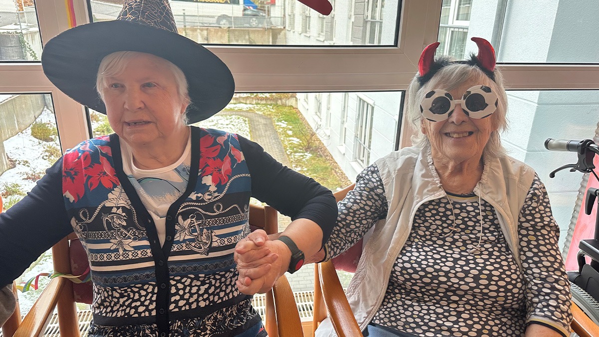 Zwei Bewohnerinnen sitzen nebeneinander, lachen und halten sich in bunten Kostümen fröhlich an den Händen.