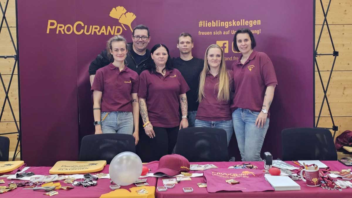The team from the care home presents itself at the trade fair stand in uniform clothing and engages in conversation with interested students. The stand is set up with informational materials and encourages discussion about the nursing profession.