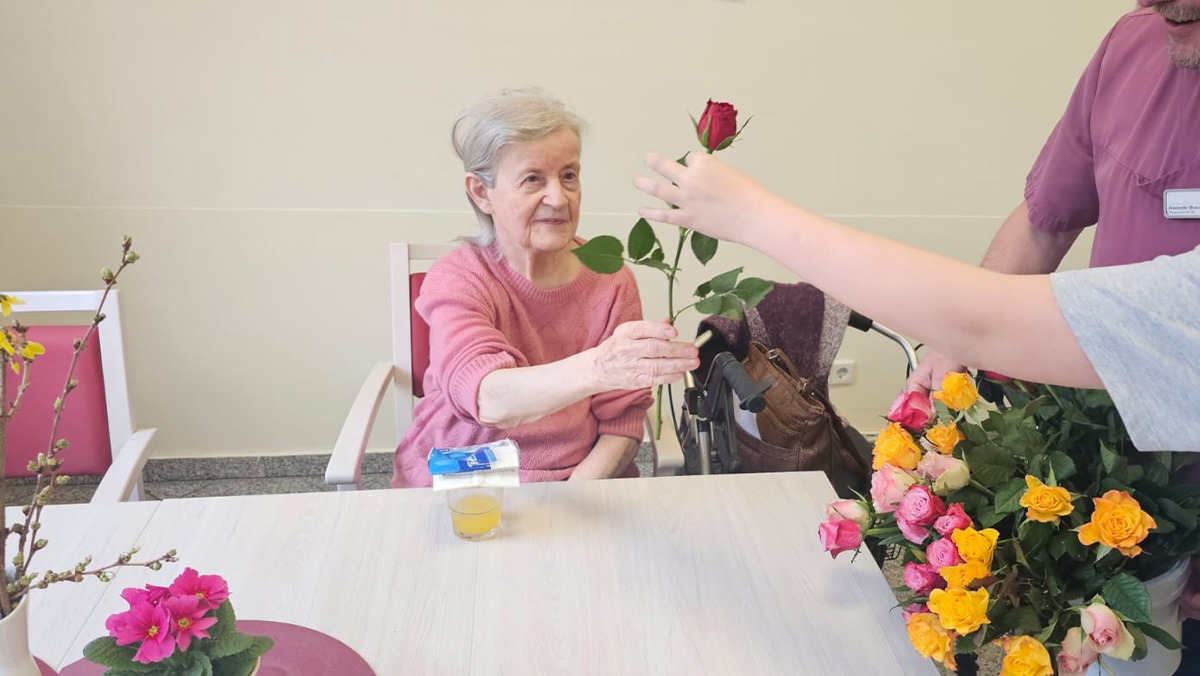 International Women's Day at ProCurand Seniorenresidenz Am Görnsee: A resident smiles as she receives a red rose from a carer. The moment radiates appreciation, joy, and care, surrounded by lovingly decorated flowers on the table.
