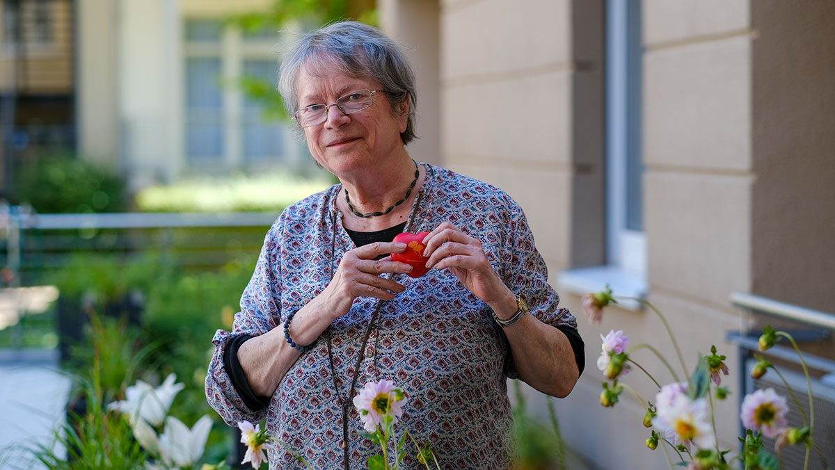 Eine Seniorin der Stationären Pflege der Seniorenresidenz Park Sanssouci steht mit einem ProCurand-Herz in ihrem Garten